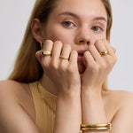 Woman wearing gold jewelry including earrings, rings, and bracelets on a plain background
